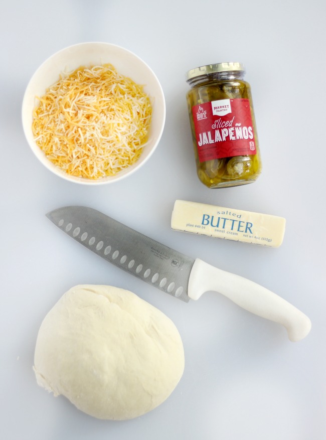 picture of a bowl of cheese, jar of japapeños, a knife, a stick of butter and a ball of bread dough