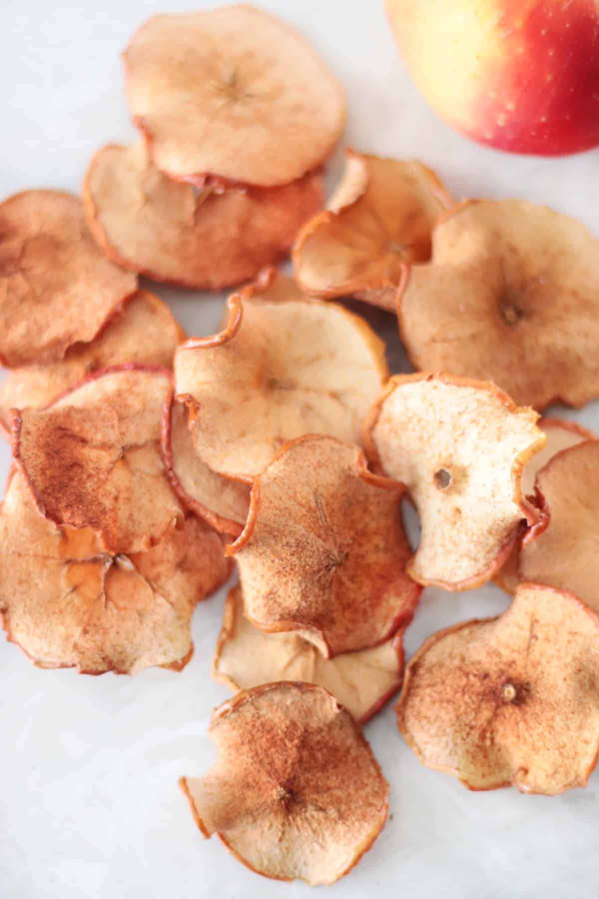 Pile of baked apple chips on table with apples in the background.