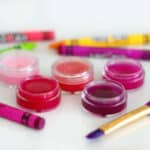 5 small containers of lip gloss next to crayons and a paint brush.