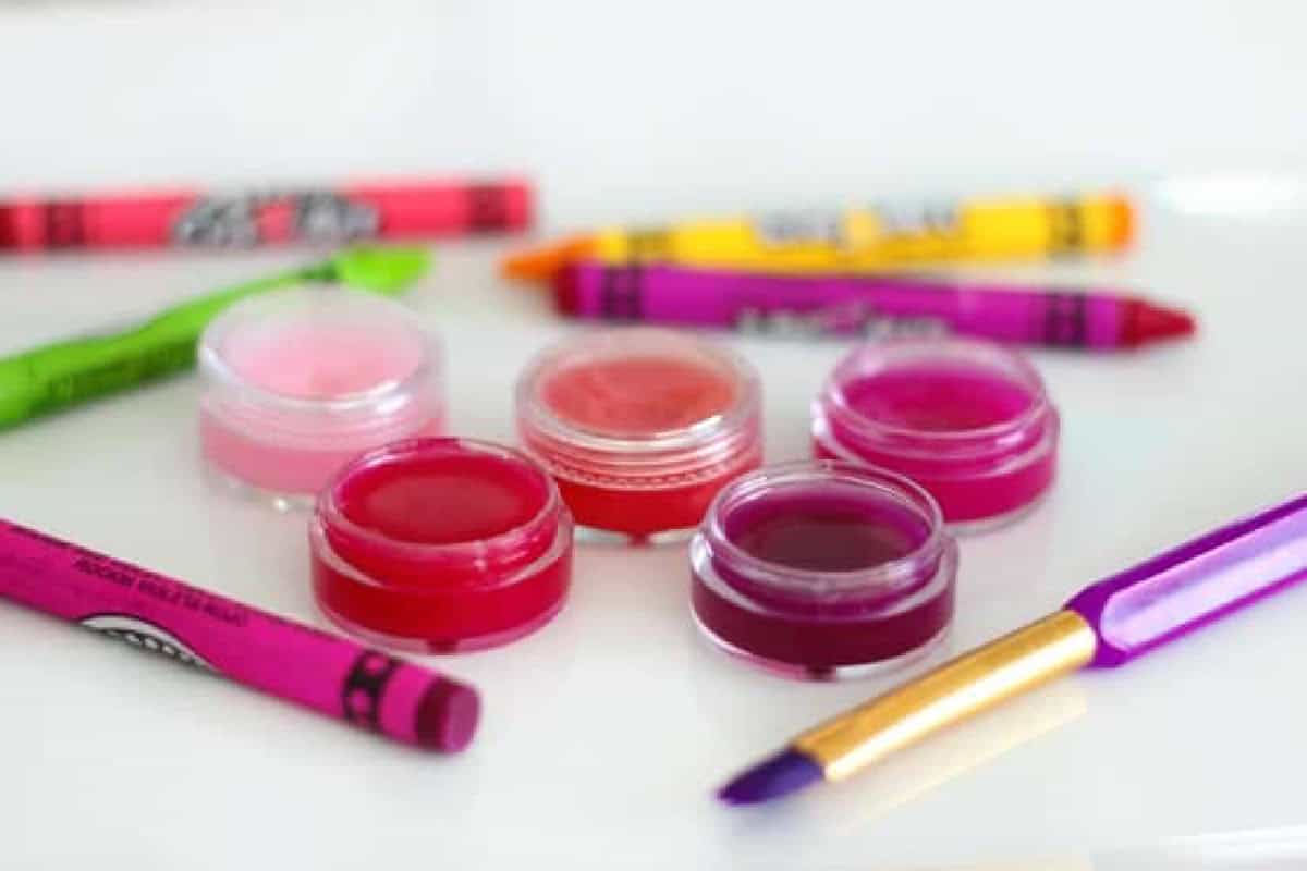 5 small containers of lip gloss next to crayons and a paint brush.