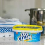 Container of oxiclean next to a stack of baby clothing and pot of water.