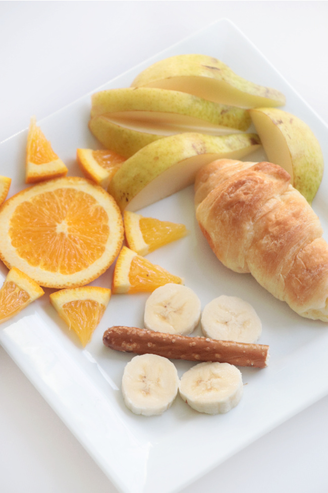 oranges, croissant, pretzel, banana slices and apples arranged on plate