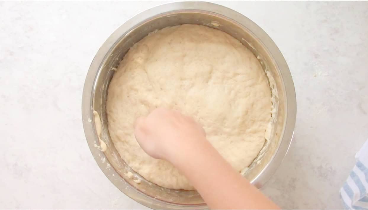 Hand punching down bread dough in large metal mixing bowl.