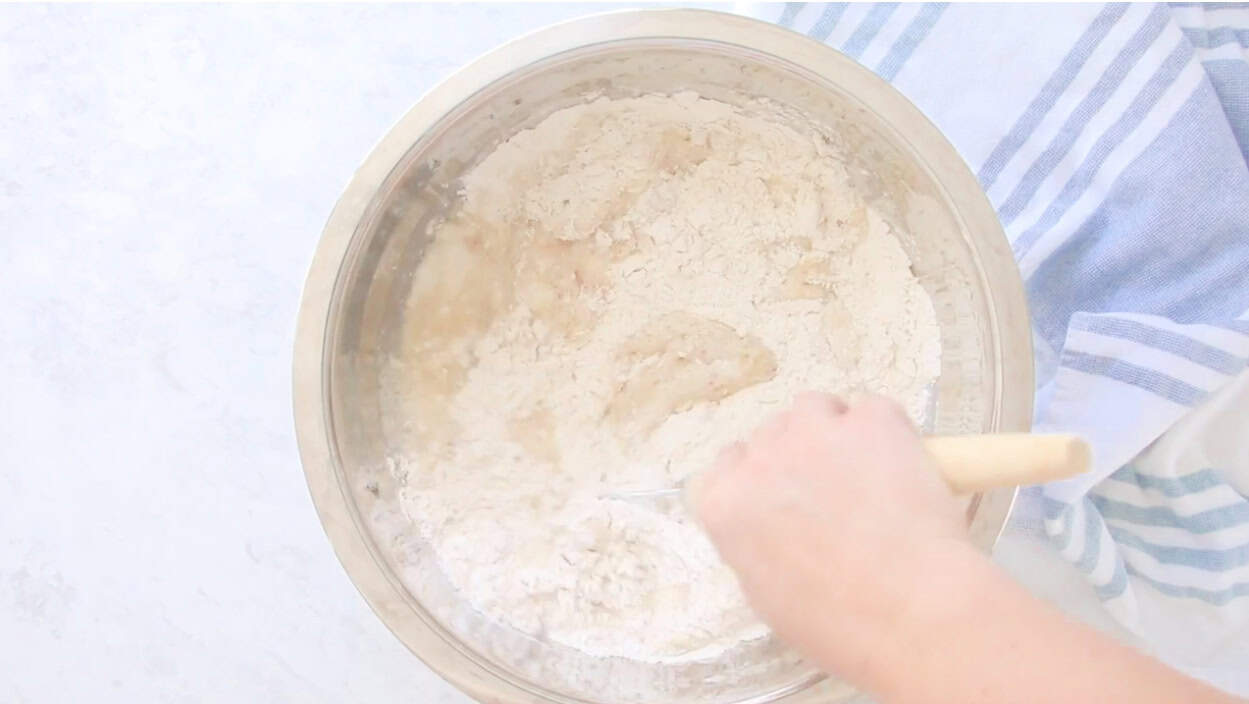 Hand stirring bread dough in large metal bowl.