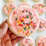 Hand holding sugar cookie with pink icing and rainbow sprinkles.