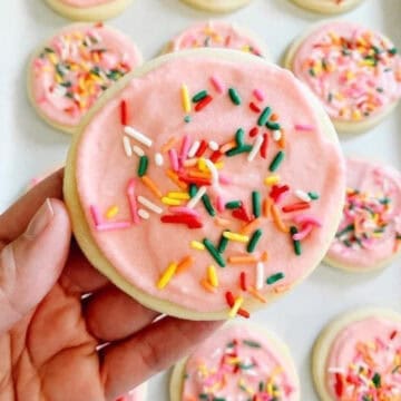 Hand holding sugar cookie with pink icing and rainbow sprinkles.