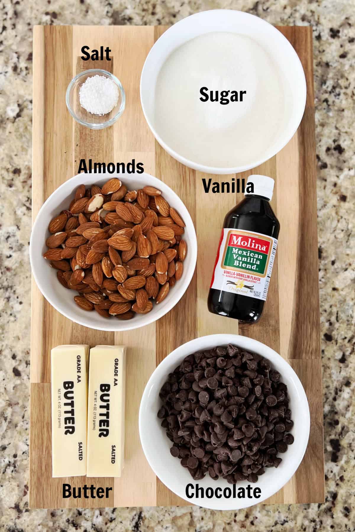 Cutting board with bowls of sugar, salt, almonds, chocolate, two sticks of butter and bottle vanilla.