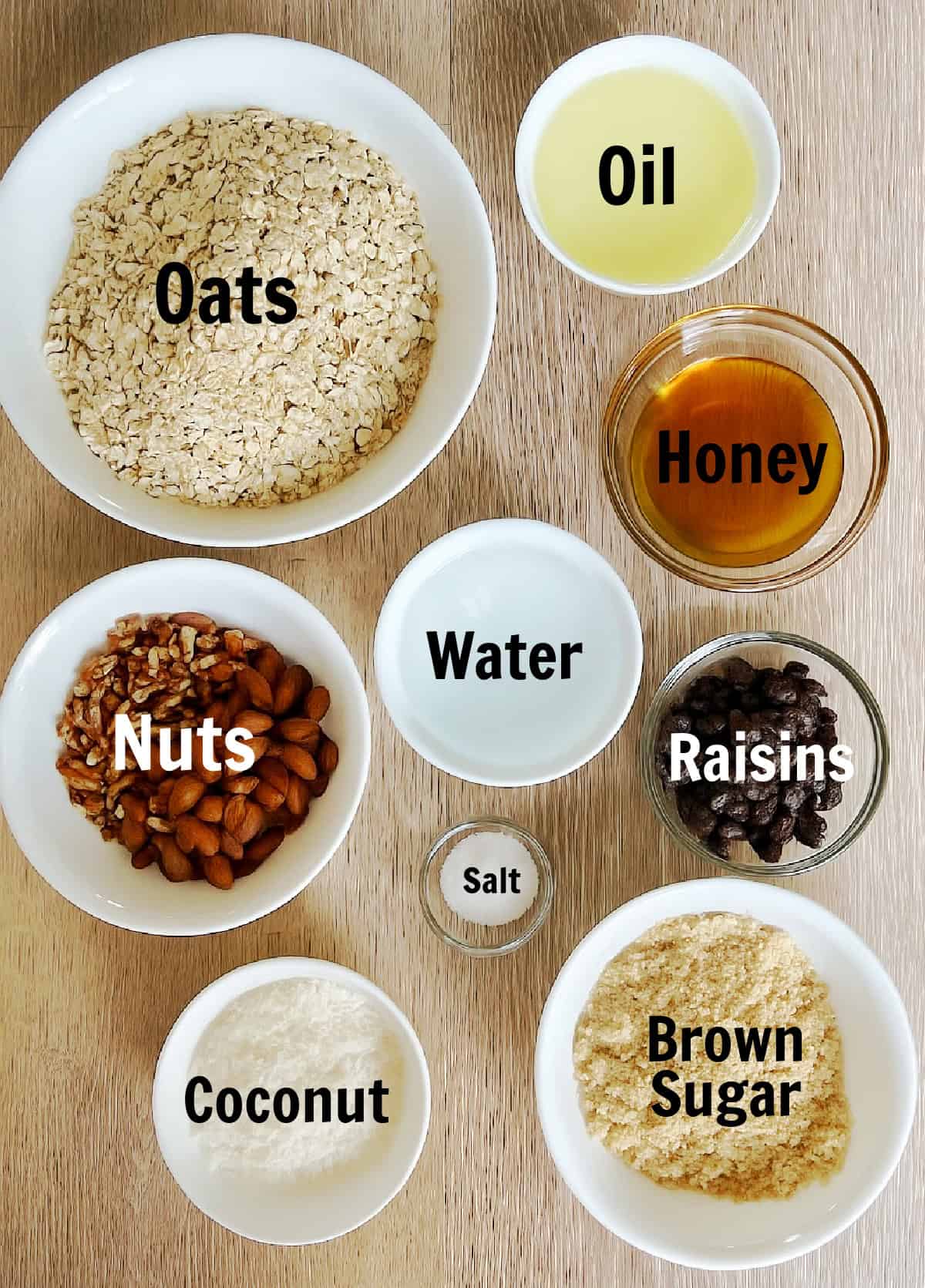 Individual bowls with oats, oil, honey, water, salt, nuts, coconut flakes, brown sugar and raisins on wooden tabletop.