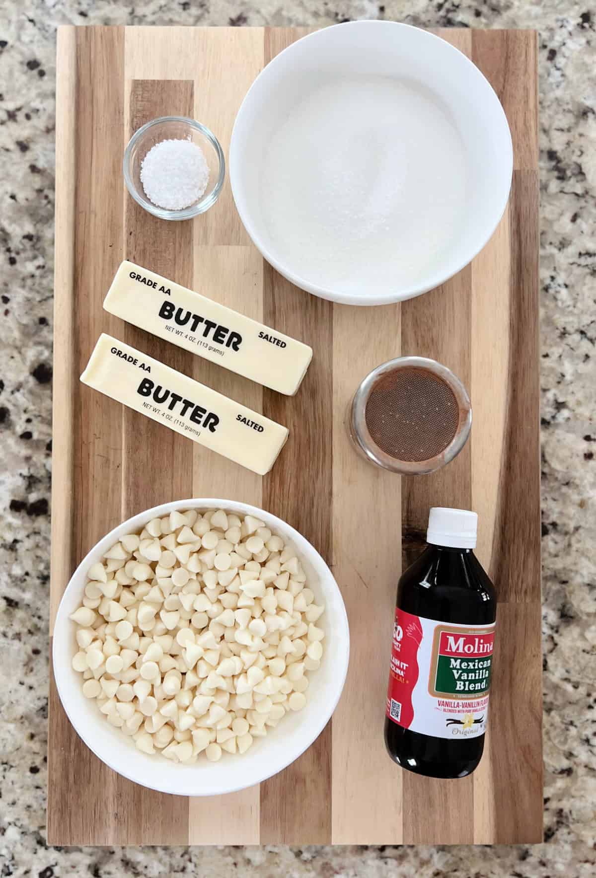 Bowls of white chocolate chips, salt and sugar. Cinnamon and sugar container, bottle of vanilla extract, and two sticks of butter.