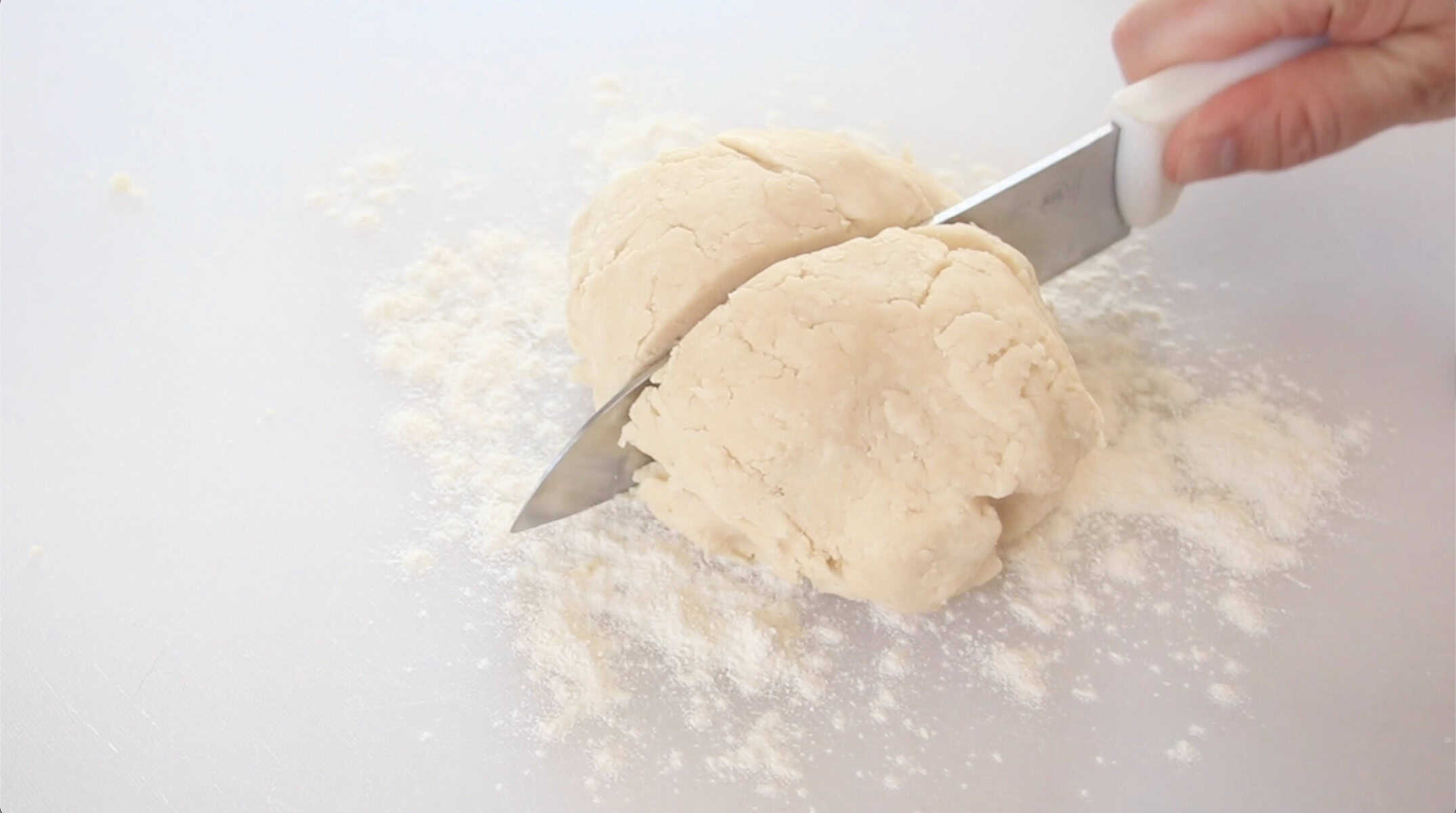 Knife slicing ball of pie dough in half on white cutting board.