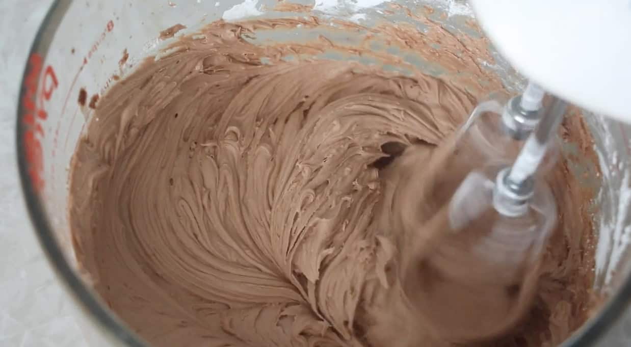 Glass mixing bowl of chocolate frosting beat with electric mixer.