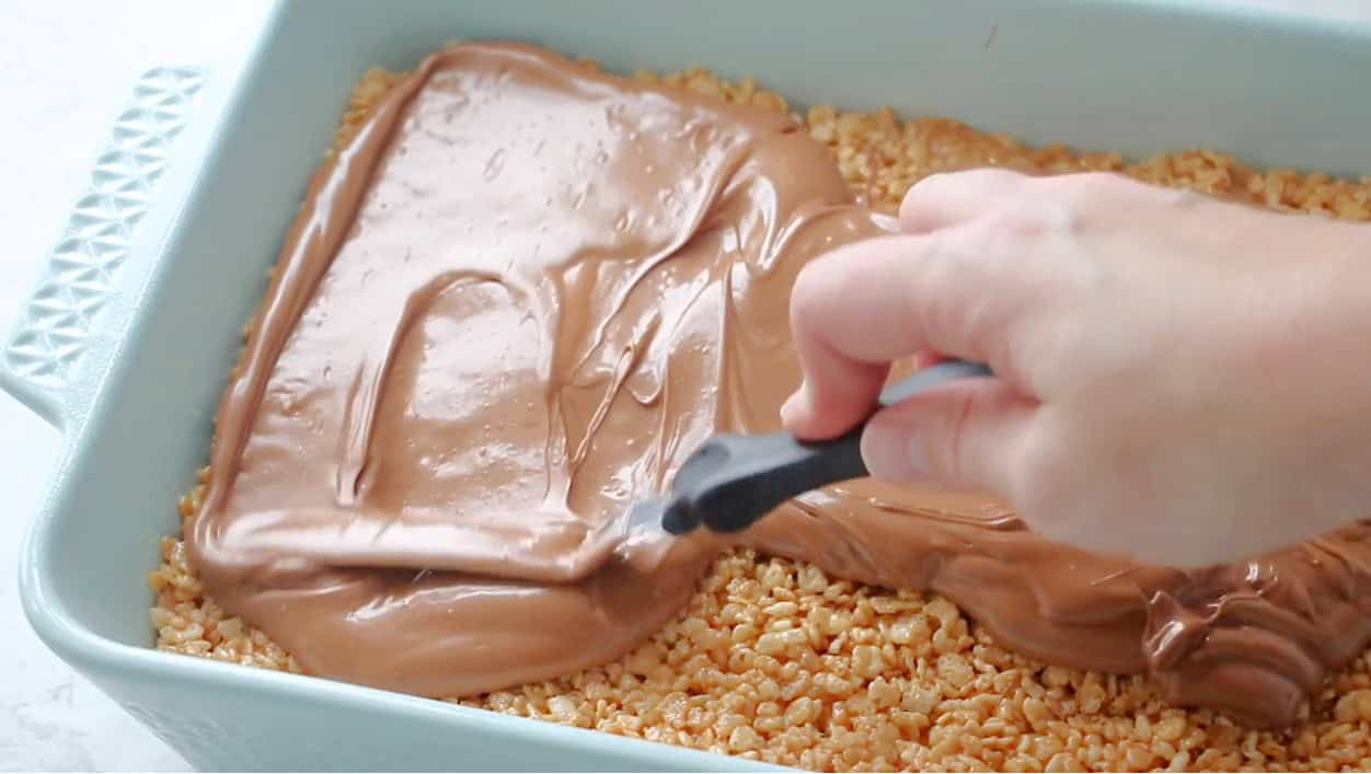 Hand using spatula to spread chocolate over scotcheroos.