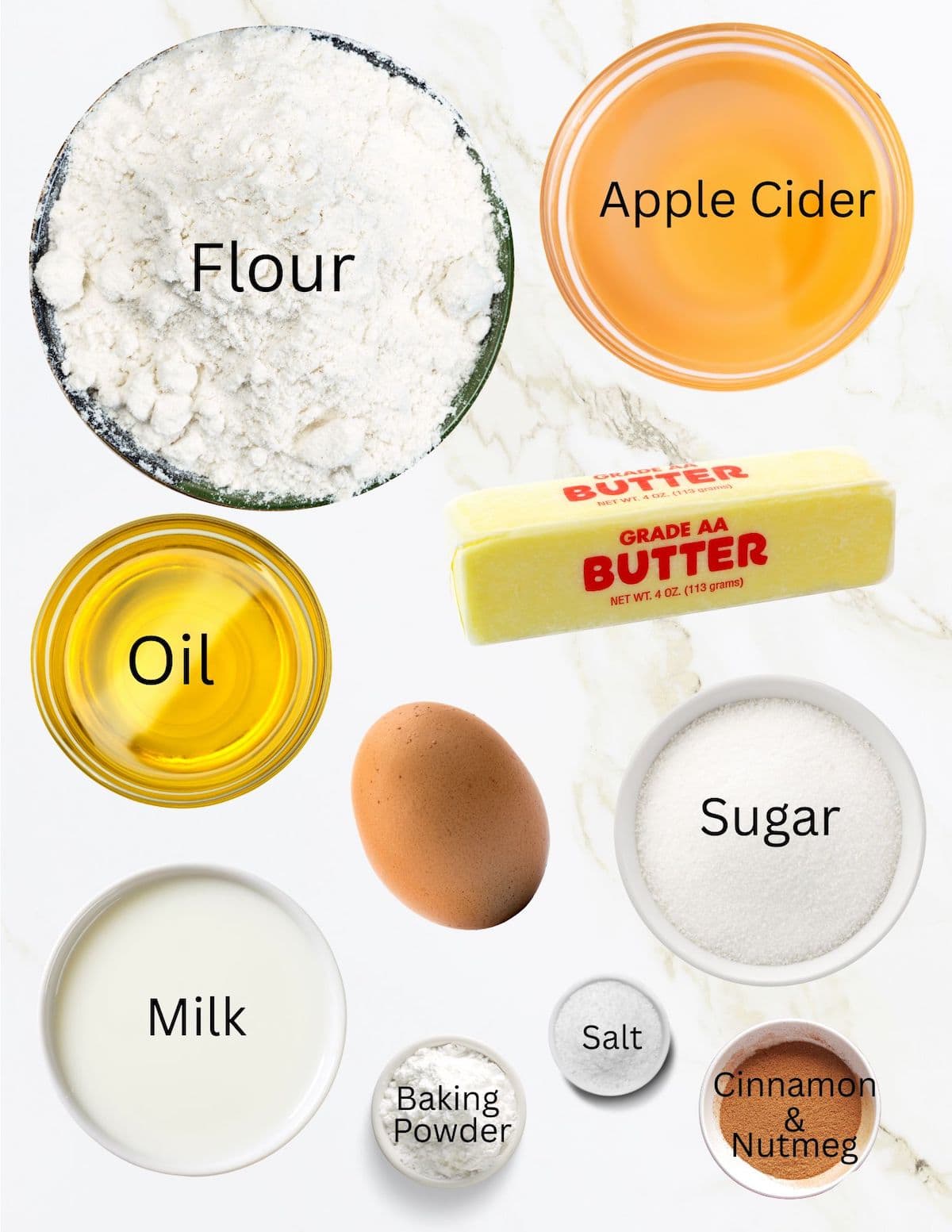 white countertop with bowls of flour, apple cider, oil, milk, sugar, baking powder, salt, cinnamon, nutmeg, stick of butter and brown egg.