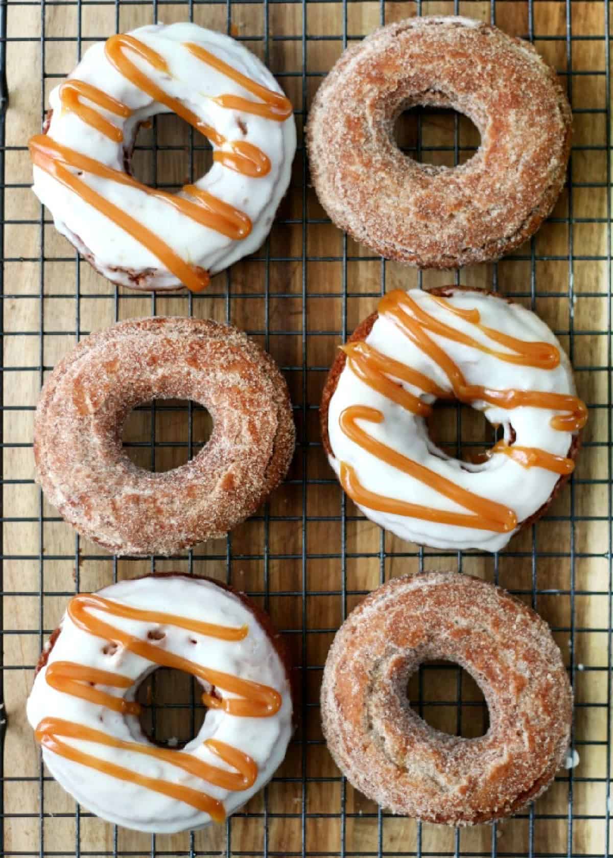 3 apple cider donuts coated in cinnamon and sugar and 3 cider dipped in buttercream and drizzled with caramel on a wire cooling rack.