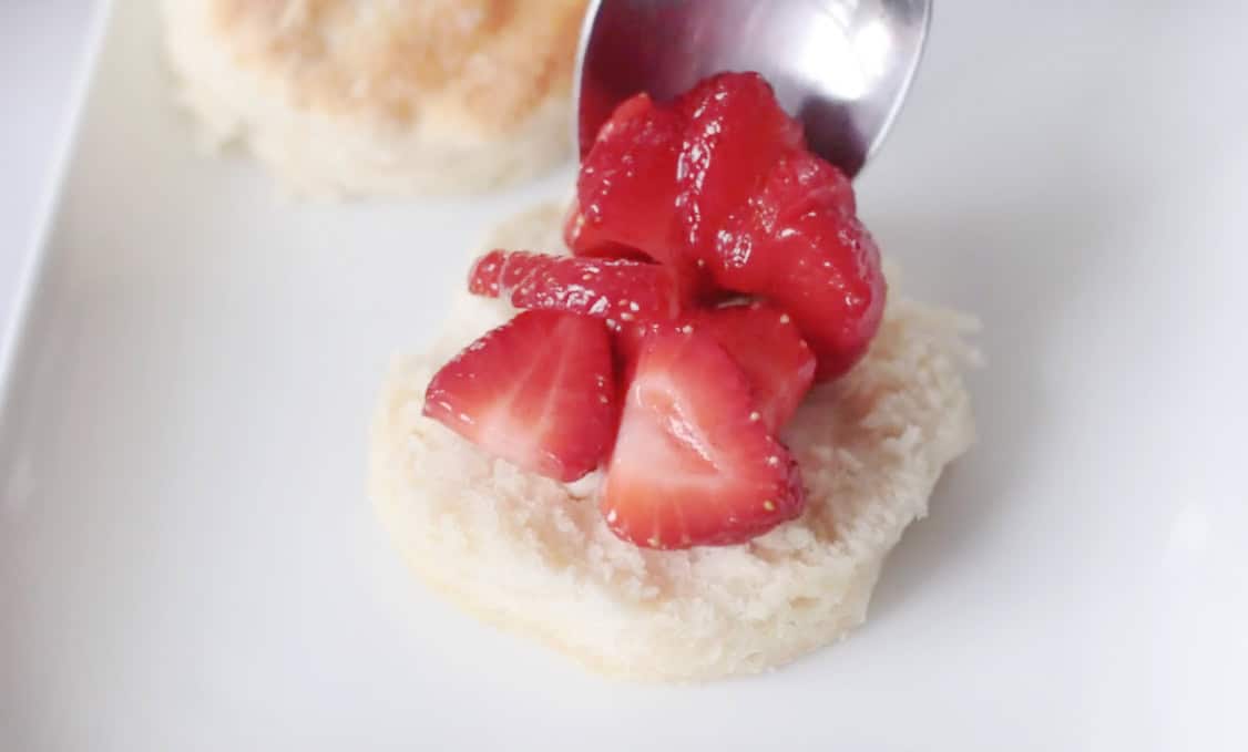 White plate with biscuit and spoon adding strawberries on top of biscuit.