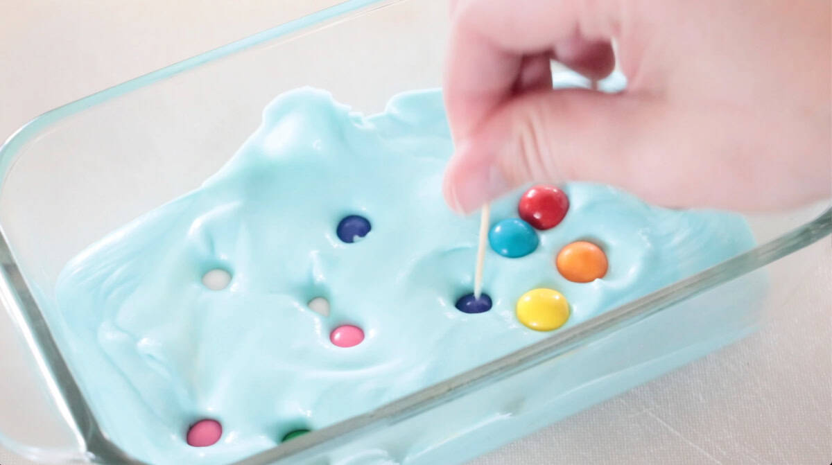 Hand using toothpick to poke and bury gum balls in ice cream mixture.