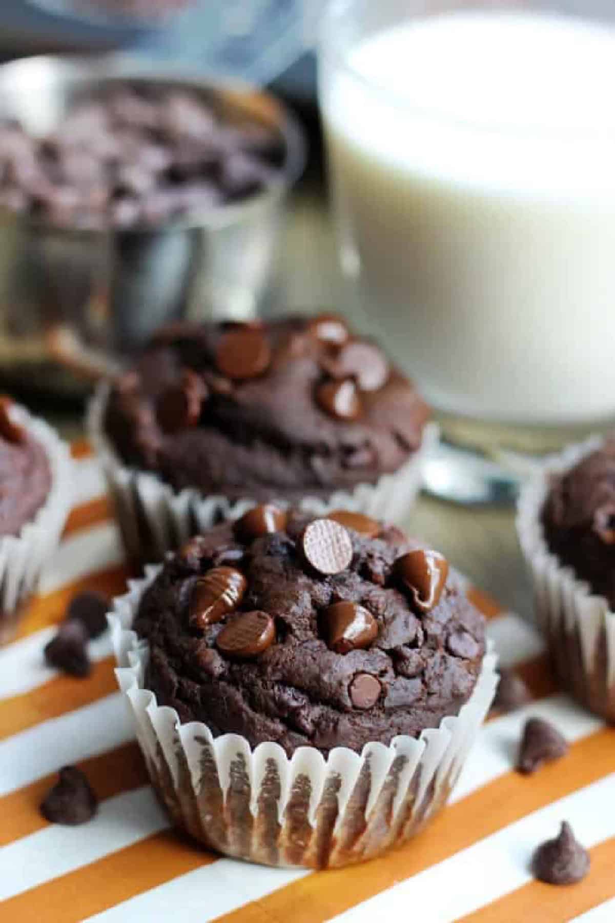 Double chocolate pumpkin muffins on orange striped napkin next to a glass of milk.