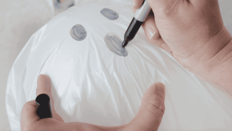 Hand holding permanent marker and drawing eyes and mouth on garbage bag ghost.