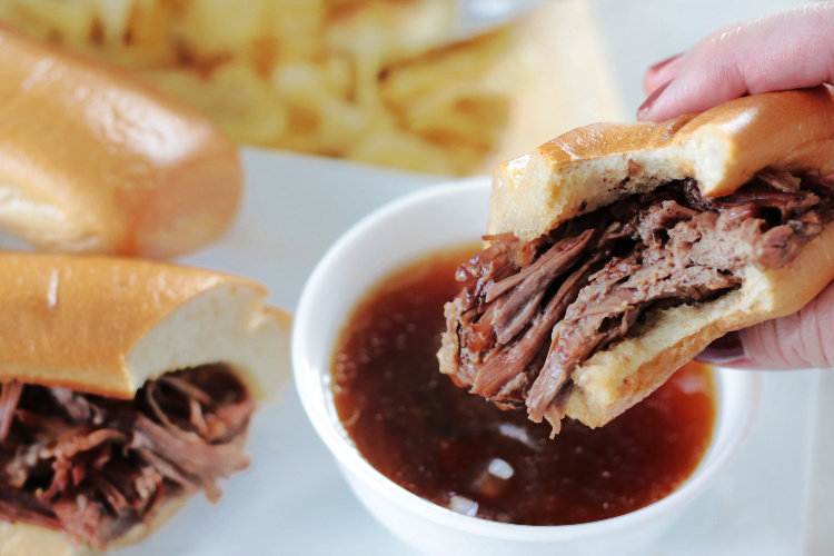 hand holding slow cooker French dip sandwich over bowl of au jus