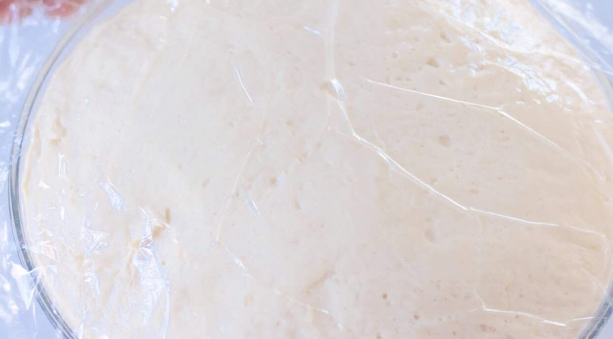 Large bowl with proofed dough and plastic wrap on top.