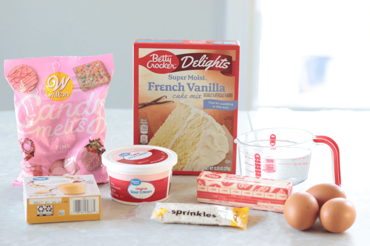 boxed cake mix, pink candy melts, box of vanilla pudding mix, sprinkles, stick of butter, container of sour cream, cup of water and 3 eggs