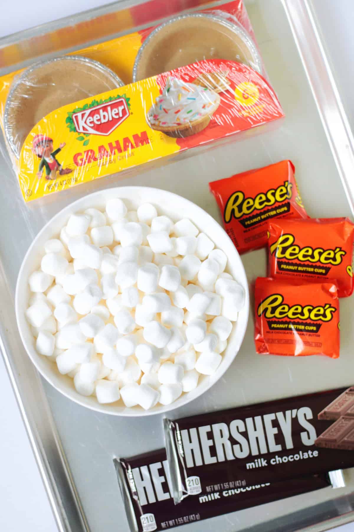 baking sheet with bowl of marshmallows, mini pie crusts, reese's peanut butter cups and hershey bars.