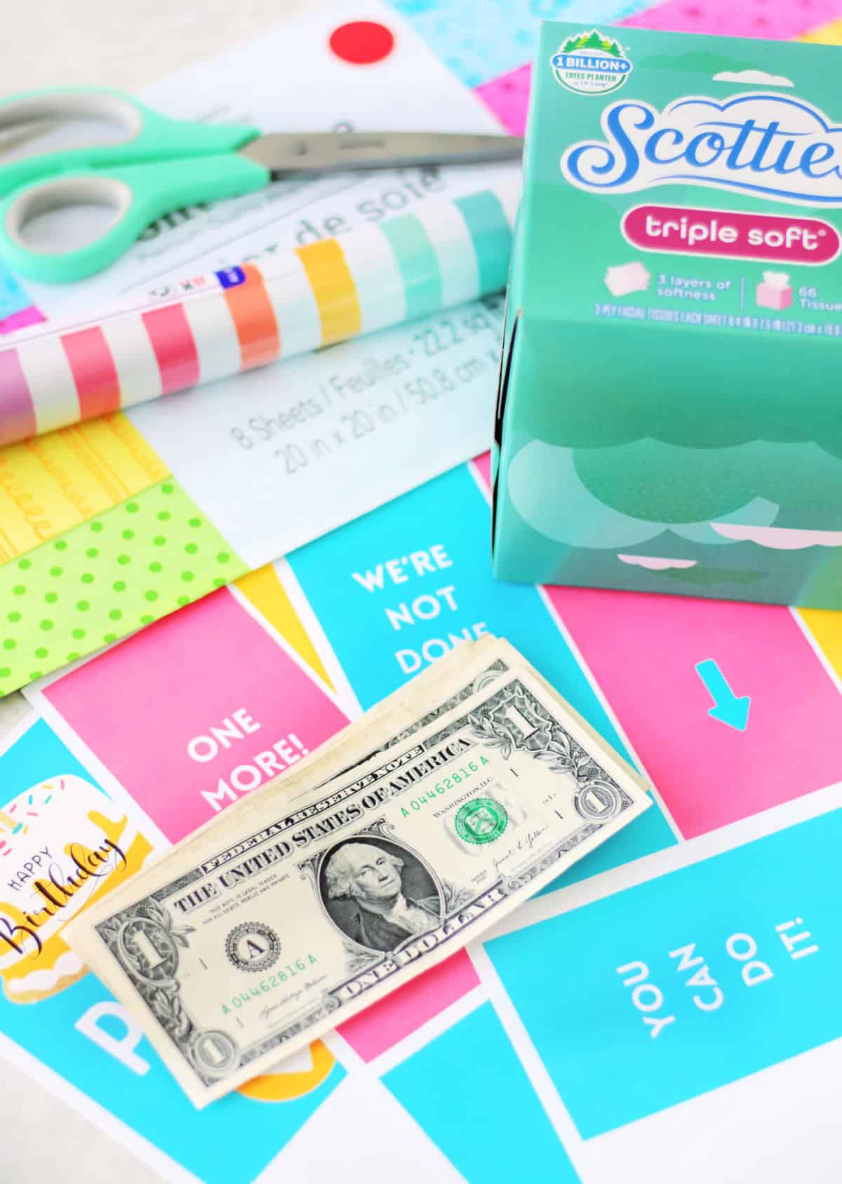 Stack of dollar bills, printable tags, tissue box and roll of wrapping paper.