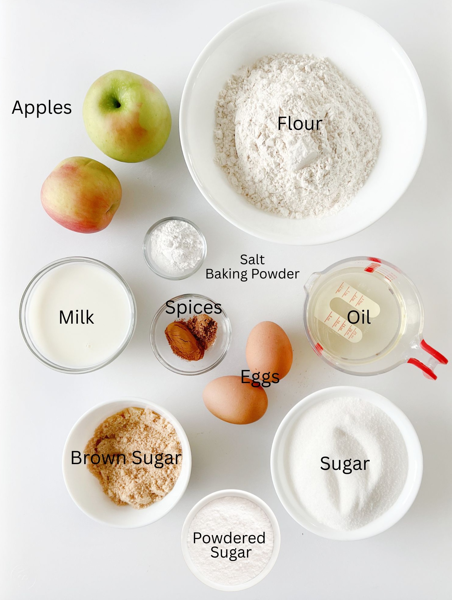 Bowls of flour, salt and baking powder, two eggs, spices, bowls of brown sugar and powdered sugar, oil, milk and apples.