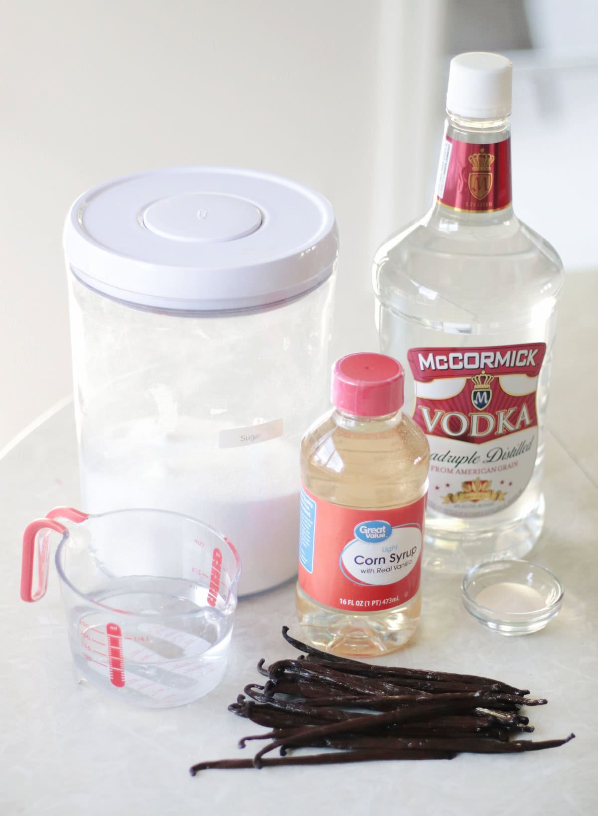 Container of sugar, bottle of vodka, cup of water, bottle of corn syrup, bowl of xanthan gum, and pile of vanilla beans.