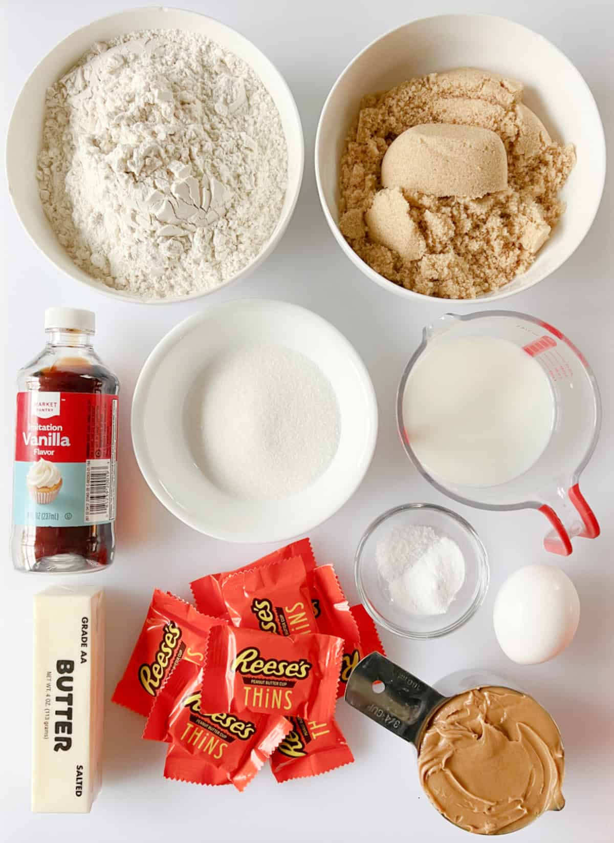 Bowls of sugar, flour, brown sugar, milk, bottle of vanilla, stick of butter, stack of peanut butter cups, cup of peanut butter, an egg and small bowl of salt, baking soda and baking powder.