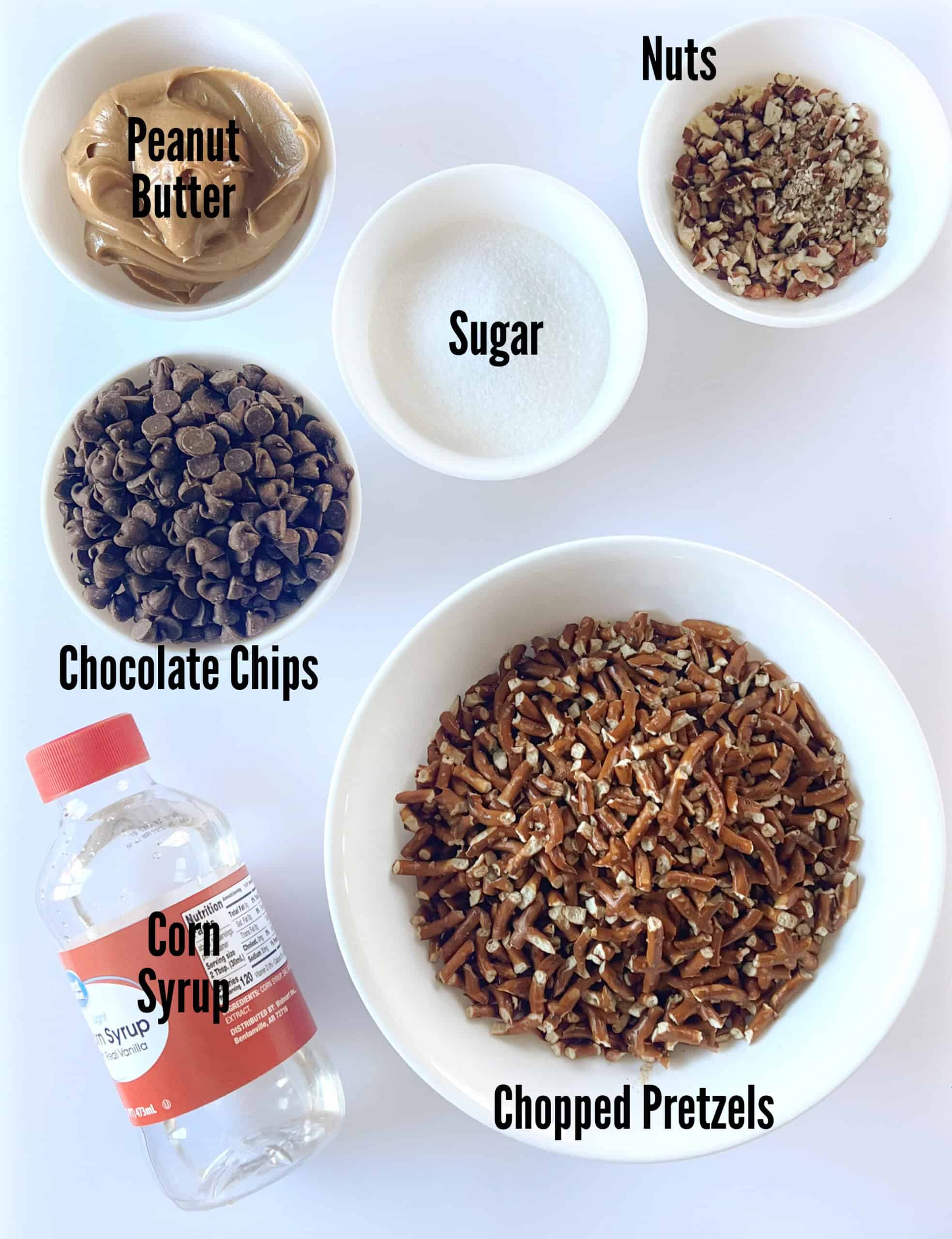 Bowls of peanut butter, sugar, pecans, chocolate chips, chopped pretzels and bottle of corn syrup.