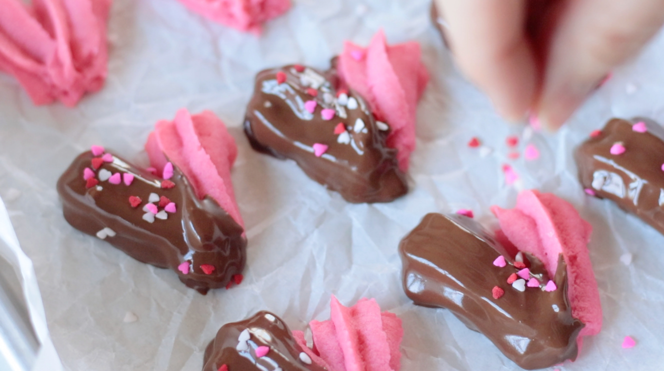 Pink heart shaped cookies dipped in melted chocolate on baking sheet.
