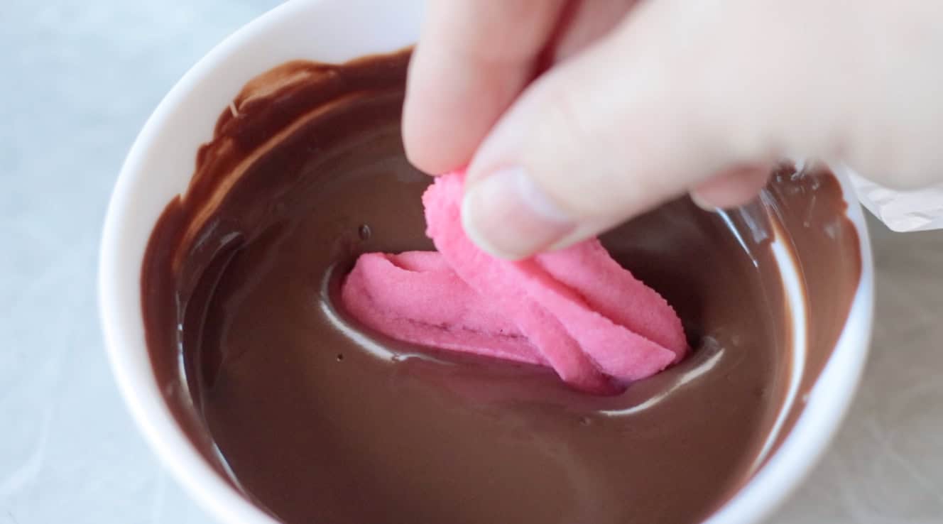 Hand dipping pink heart butter cookie in melting chocolate.