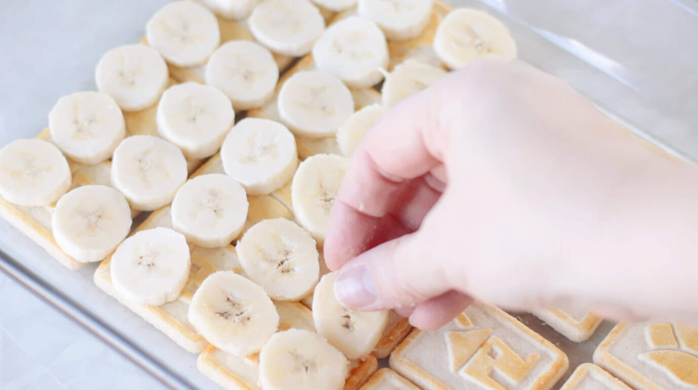 Hand placing slices of banana on top of vanilla pudding in baking dish.