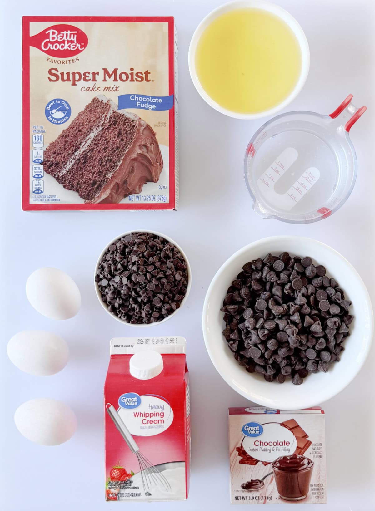 Box of chocolate cake mix, bowl of oil, bowl of mini chocolate chips and regular chocolate chips, container of cream, 3 eggs, box of chocolate pudding mix, and cup of water.