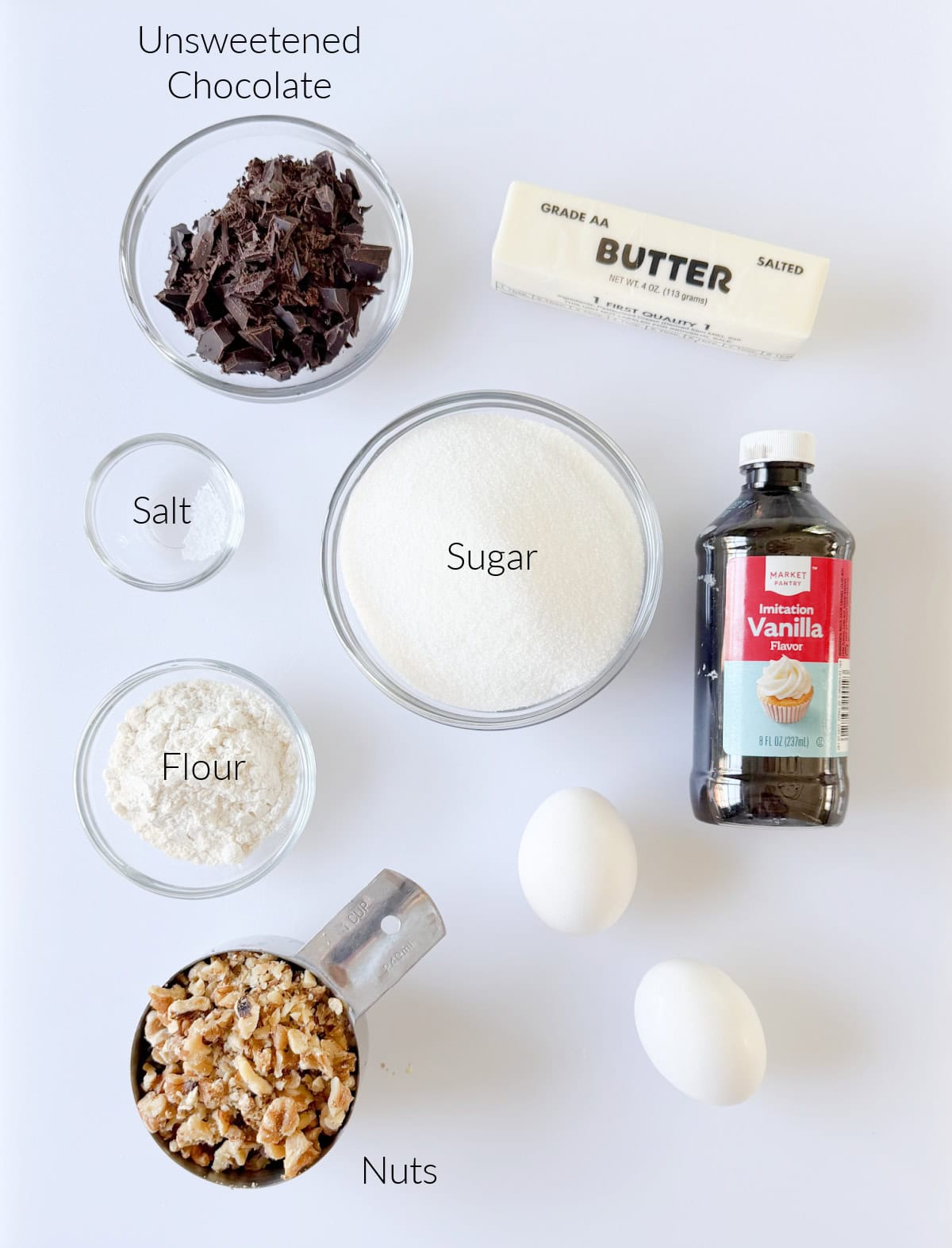 Bowl of chocolate, stick of butter, bottle of vanilla, bowls of sugar, salt and flour, measuring cup of walnuts and two eggs.