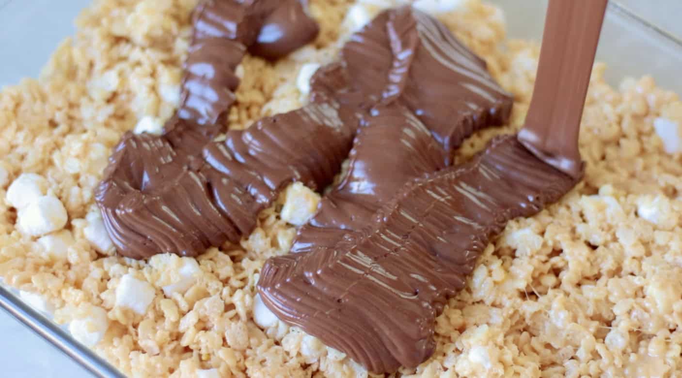 Baking dish with pressed scotcheroo mixture and chocolate drizzled over the top.