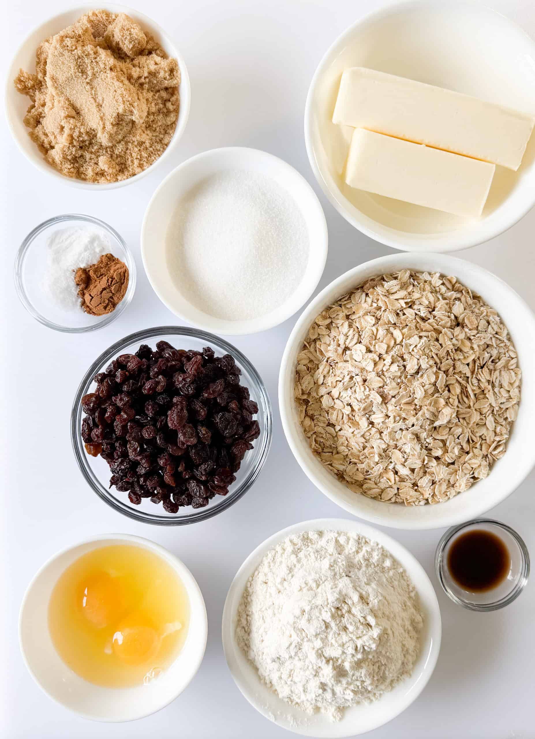 Bowls of brown sugar, white sugar, butter, spices, raisins, oats, eggs, flour and vanilla extract.