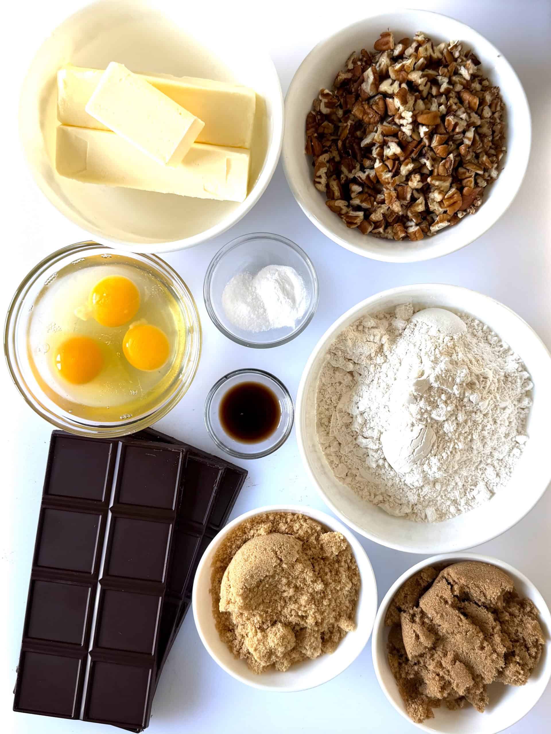 Bowls of butter, pecans, eggs, baking soda and salt, flour, dark brown sugar, light brown sugar, baking chocolate bars.