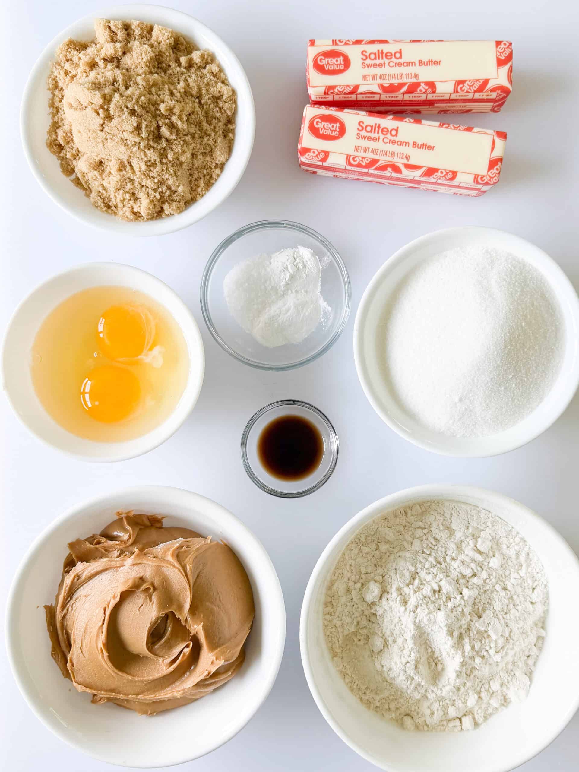 Bowls of brown sugar, eggs, white sugar, baking soda, baking powder and salt, vanilla, flour, peanut butter and two sticks of butter.