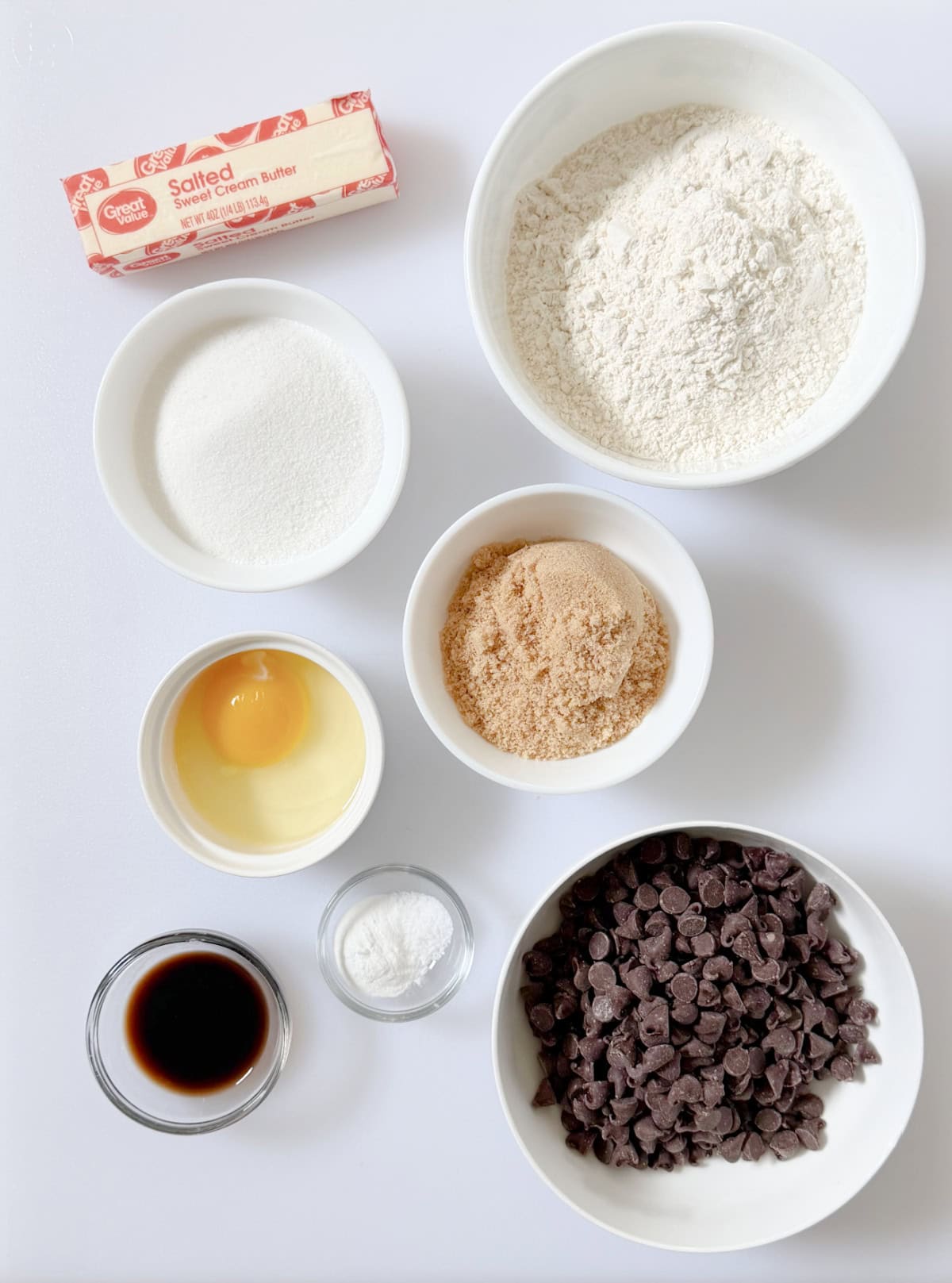Stick of butter, bowls of sugar, brown sugar, flour, egg, vanilla, chocolate chips and baking soda and salt.