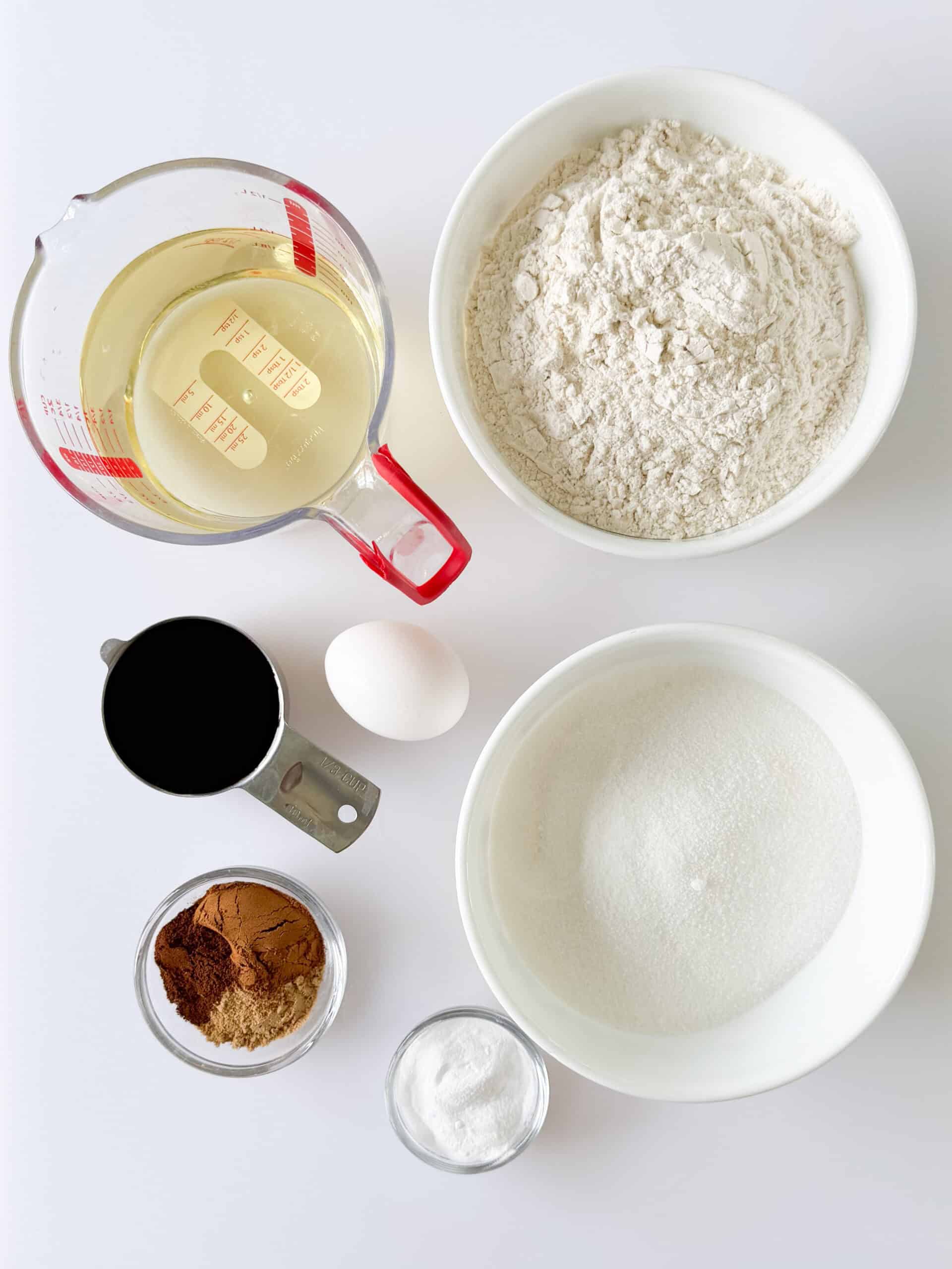 Bowls of flour, sugar, spices, baking soda, an egg, measuring cup of oil and measuring up of molasses.