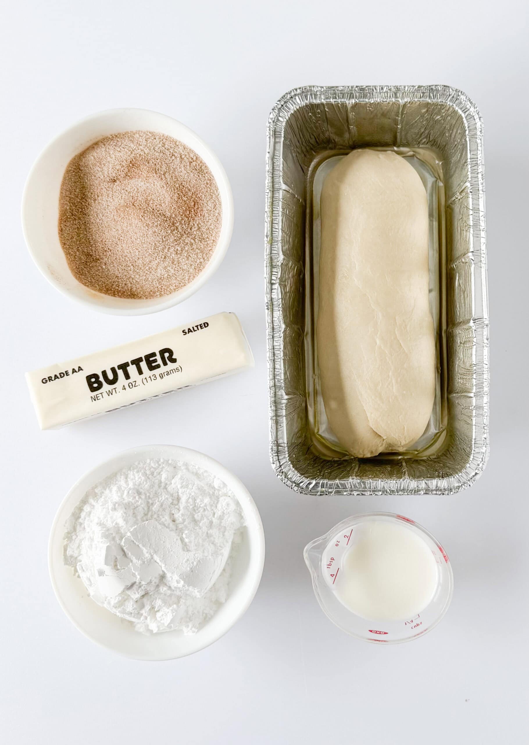 Bowl of cinnamon and sugar, loaf of frozen bread dough, bowl of powdered sugar, stick of butter and small bowl of milk.