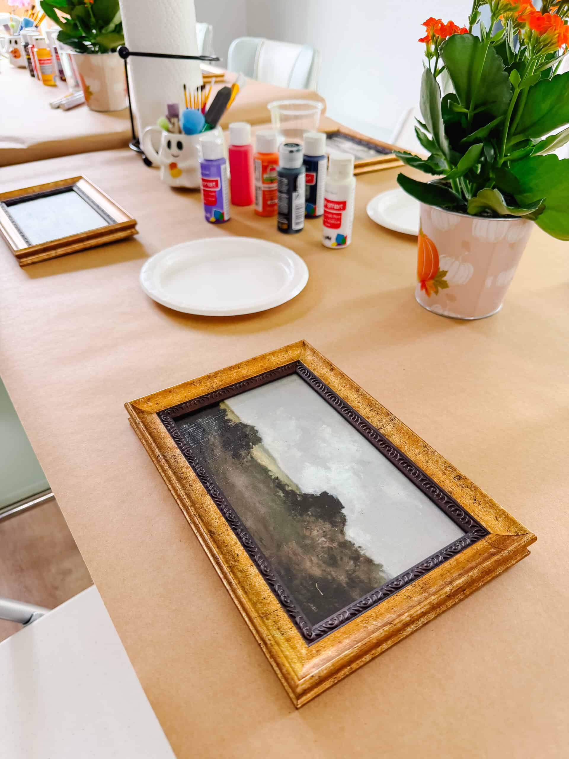 Framed artwork on table with bottles of paint, paper plates and paint brushes.