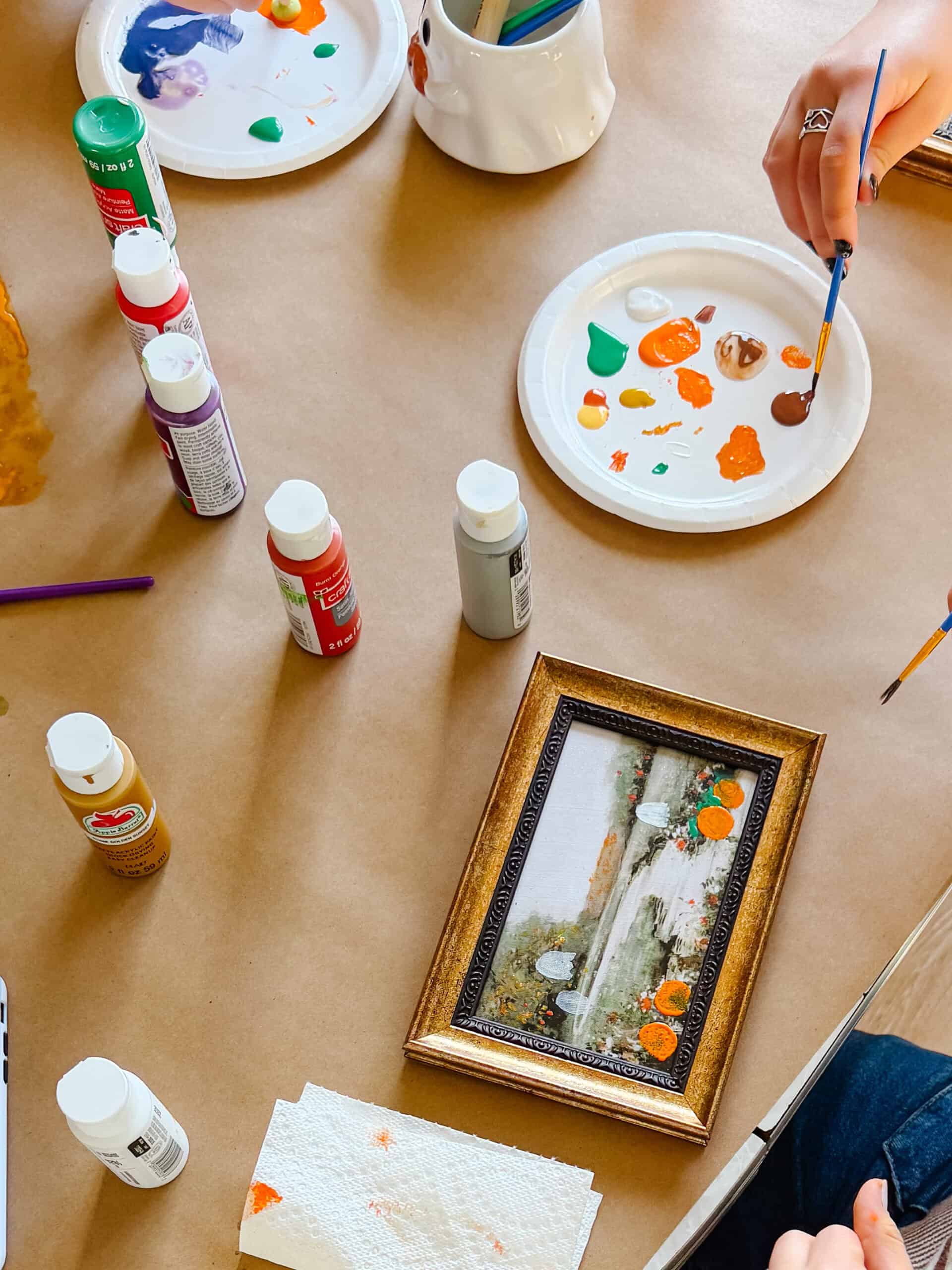Paper plate of paint and bottles of acrylic paint next to framed artwork being painted on.